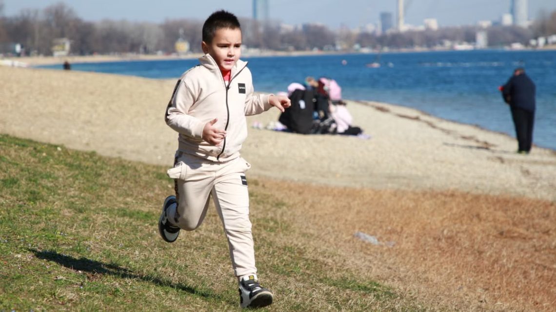 Young boy in a tracksuit runs energetically by the waterfront, embodying the joy of a sunny day outdoors.